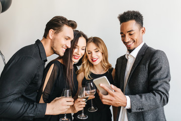 Pleased african man in vintage tweed jacket making selfie with friends at birthday party. Indoor portrait of black guy holding smartphone and charming girls drinking champagne on event.