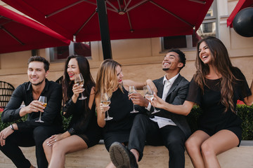Young woman with white manicure holding glass of wine and laughing with husband. Portrait of chilling friends celebrating vacation in outdoor cafe and talking around.