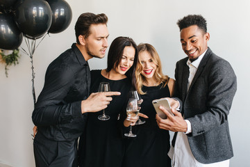 Joyful african man making funny face posing at birthday party with gorgeous girls. Attractive ladies with wineglasses smiling on white background while black guy taking selfie.