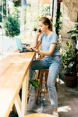 Beautiful blonde girl with a laptop in a cafe by the window talking on the phone