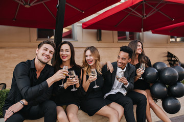 Laughing guy with short dark hair clink glasses with his girlfriend during rest in cafe. Smiling friends enjoying champagne after workday, chilling in outdoor restaurant.