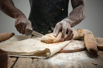 Close up of african-american man slices fresh cereal, white bread, bran with a kitchen knife on wooden table. Healthy eating, nutrition, craft product. Gluten-free food, vegan lifestyle, organic taste