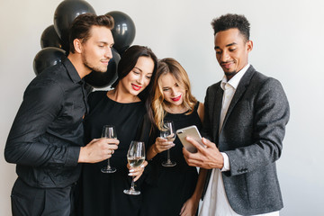 Confident african man in stylish gray jacket making selfie on event with colleagues. Pretty blonde girl looks with interest at phone screen during birthday party with best friends.