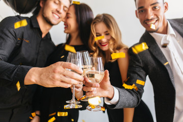 Blur portrait of magnificent curly birthday girl enjoying holiday with friends. Photo of excited young people standing under confetti with glasses of champagne in foreground.