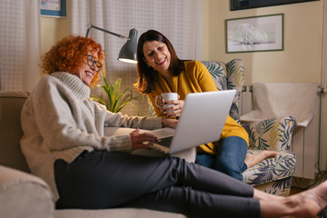 middle aged female friends having fun at home, drinking coffee and using laptop computer