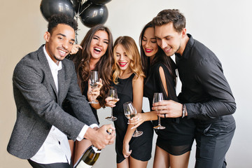 Handsome african man with beard opens bottle of champagne to celebrate something with friends. Indoor portrait of blissful young people spending time together at birthday party.