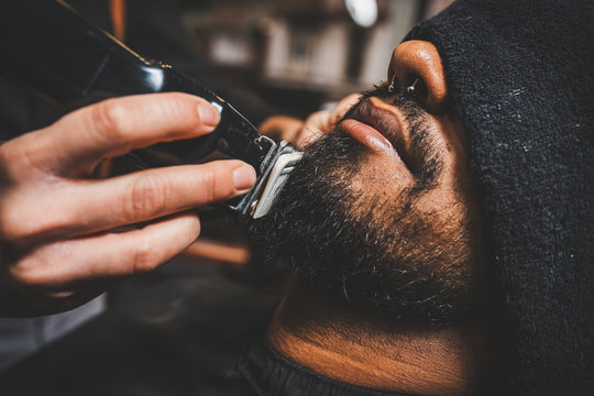 un barbero está pasando la maquina eléctrica de cortar y afeitar por la barba de un chico brasileño afroamericano. de estilo swing hipster.