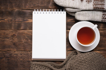Obraz na płótnie Canvas Blank notepad with copy space, warm clothes and cup of tea on brown wooden table flat lay background.