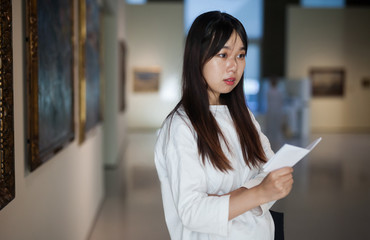Fototapeta na wymiar Chinese female visitor with guide-book looking at artwork painting
