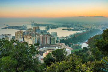 Obraz premium Evening sun over the Spanish city of Malaga on the Mediterranean coast. View of the city center on the Costa del Sol with illuminated buildings, trees, street lamps, harbor, ships