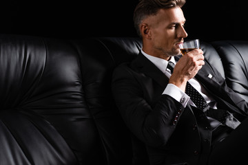 Handsome businessman drinking whiskey while sitting on couch isolated on black