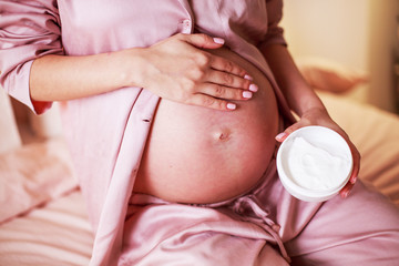 Taking care. Pregnant woman spreading cream on her belly in her bedroom.