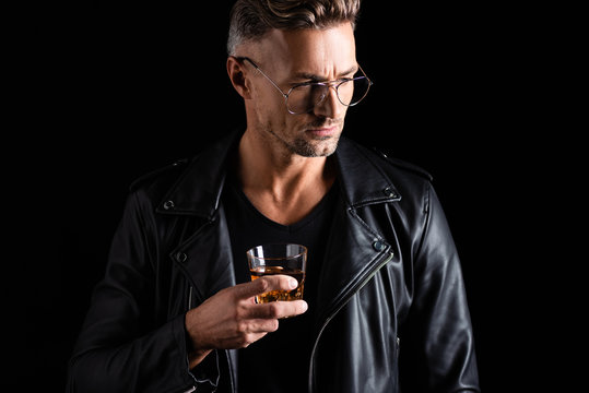 Stylish man in biker jacket and sunglasses holding glass of whiskey isolated on black