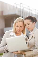 Young serious businesswomen working on laptop while standing against office building