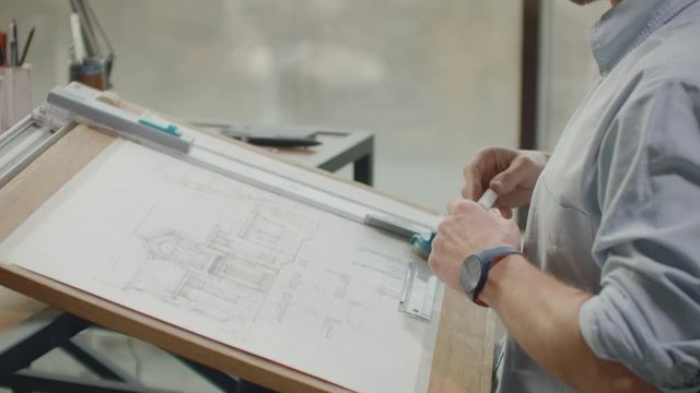 A male architect sitting at a table draws a plan of the building and is engaged in design development, sitting in the office in the Sitel loft near a large window