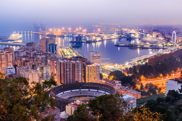 Naklejka premium Night shot of Malaga on the Spanish Mediterranean coast. City view on the Costa del Sol with illuminated harbor, residential buildings, trees, street lamps, ferris wheel, ships