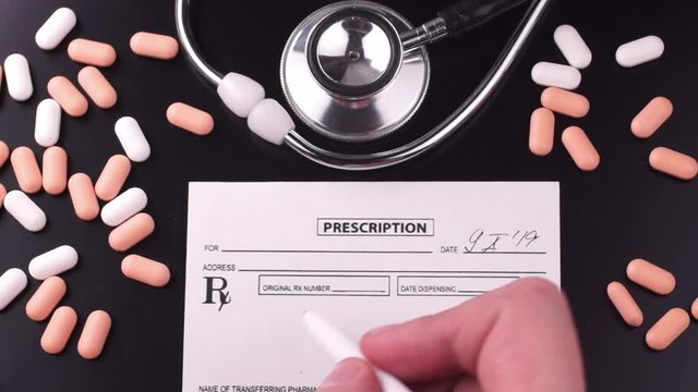 Physician fills a prescription with a white pen on a dark background next to phonendoscope and pills. Flat lay