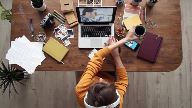 Flat lay of young woman with headphones on head looking through photos on laptop screen, on phone, making notes in notebook while having bite