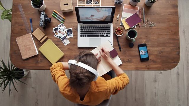 Time lapse of young female photographer sitting at laptop, putting headphones on head while looking through photos on laptop, on telephone, writing in notebook and having bite