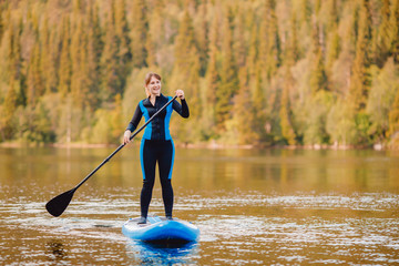 Girl in thermo clothing rowing oar on sup board blue lake water paddleboard background of forest