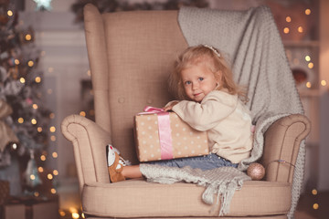 Fototapeta premium Funny baby girl 3-4 year old open Christmas present sitting in chair over xmas lights closeup in room. Looking at camera. New Year. Winter holiday season.