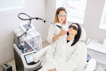 Charming female beautician does ultrasonic face cleaning pretty woman client sitting in a chair in a white coat and safety glasses in beauty spa clinic. ?oncept of skin cleansing procedures