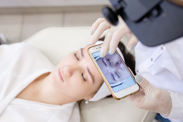 Unknown beautician's hands in gloves take photos on smartphone of the client's girl to use the program to select the perfect eyebrow shape. Microblading and eyebrow tattoo concept.