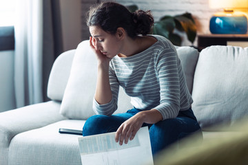 Worried young woman thinking about her problems while holding bills of home.
