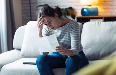 Worried young woman looking at bills and thinking about her problems while sitting on the sofa at...