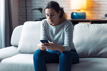 Serious young woman using her mobile phone while sitting on sofa in the living room at home.