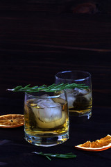 two glasses of whiskey and ice on wooden table. glass of lemonade with pieces of ice and a sprig of rosemary with slices of orange on a dark background.