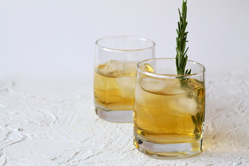 glass of lemonade with pieces of ice and a sprig of rosemary on a white background. two glasses of whiskey and ice on white table. horizontal