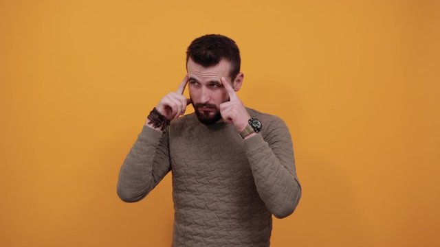Handsome caucasian cheerful man over isolated orange background keeping fingers on head, thinking about issue wearing casual green sweater