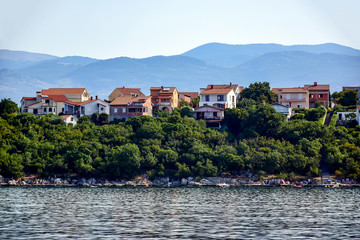 Krk, Croatia - Houses with brown roofs on a hill with trees, near the Adriatic Sea in the background of mountains, in the summer afternoon.