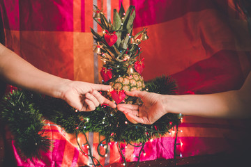 Woman's and man's hands decorating pineapple as alternative Crhistmas tree, with ornaments and Christmas lights on colorful background