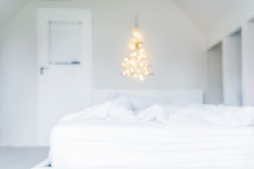 Blurred background of glowing lights Christmas tree on the white wall above the bed in the bedroom. Simple, minimal conscientious Christmass interior design.