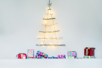 Modern creative christmas eco tree made of wooden sticks hanging on white wall with festive lights and colourful gift boxes. Simple, minimal conscientious interior design and decor. Copy space.