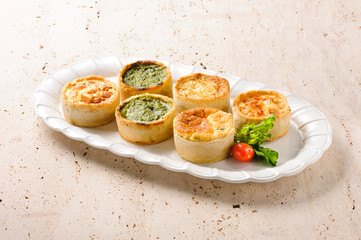 Six different and saborous quiches, on top of a white serving dish on a stone table