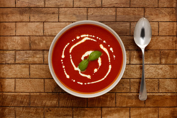 A bowl of delicious tomato soup, topped with a drizzle of cream and a sprig of basil, in a rustic bowl, shot on a wooden background, with a spoon.
