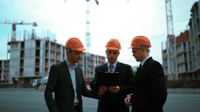 4k. Ultra HD. businessmen and architect sign document on textpad. They happy of good deal, smile and speak. Orange helmets black suit and tie. Static middle shot with modern building bg. Crane and bea