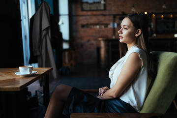 a girl sits at a cafe table, a cup of coffee is waiting for her, and she looks out the window