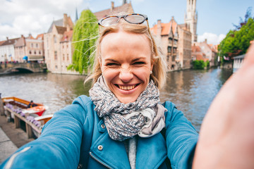 Fototapeta na wymiar Beautiful young girl takes selfie photo on the background of the famous tourist destination with a canal in Bruges, Belgium