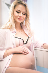 Young beautiful woman with a pregnant belly stroke her stomach, rejoices to her newborn child sitting in a shirt and underwear in a beautiful interior