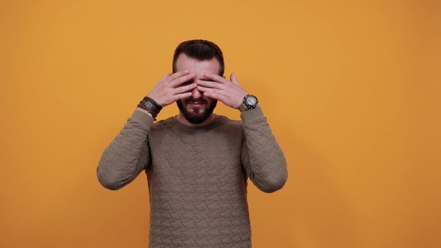 Smiling handsome caucasian man over isolated orange background looking at camera, covering eyes, looking through finger, wearing fashion green sweater