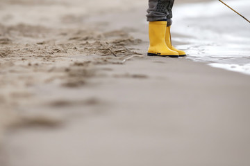 Kid in bright yellow rubber boots is standing at the surf zone of a sandy sea shore - foreground...