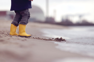 Kid in bright yellow rubber boots is standing at the surf zone of a sandy sea shore in front of a...