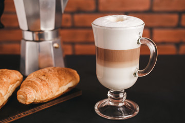 Glass with latte macchiato coffee, croissant and moka pot at dark table on brick wall background