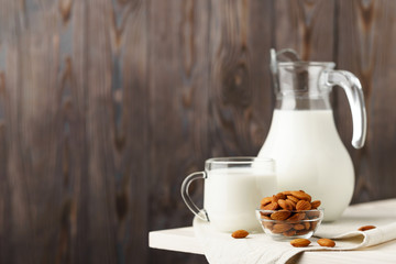 Almond milk in a glass jug and mug, next to nuts in a bowl and a textile napkin on the edge of a white wooden table, wood background.