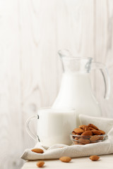 Almond milk in a glass jug and mug, next to nuts in a bowl and a textile napkin on the edge of a white wooden table, wood background.