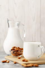 Almond milk in a glass jug and mug, next to nuts in a bowl on a concrete background.